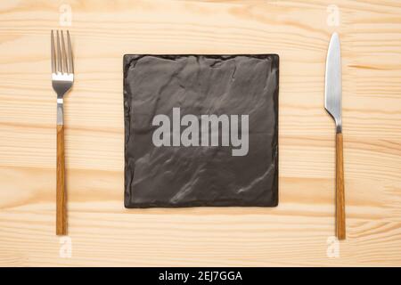 Knife and fork with square black slate board for menu or recipe text on black table. Mockup Blank board and cutlery on wooden rustic table background Stock Photo