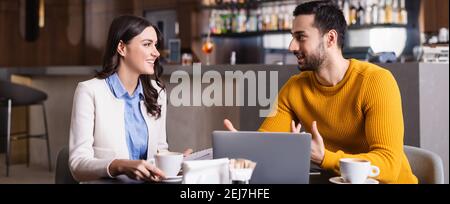 arabian freelancer talking to smiling friend near laptop in restaurant, blurred foreground, banner Stock Photo