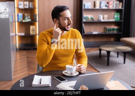 arabian freelancer adjusting earphone near laptop and cup of coffee in restaurant Stock Photo