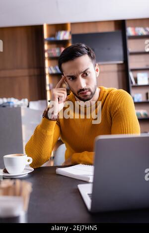 thoughtful arabian freelancer looking at laptop while working in restaurant, blurred foreground Stock Photo