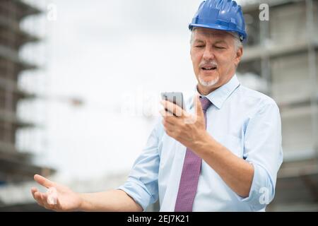 Angry and stressed architect looking at his mobile phone in front of a construction site Stock Photo