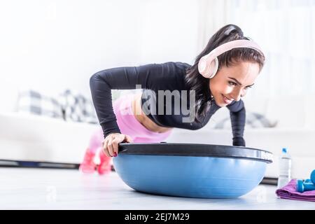 Beautiful fit woman does workout at home with headphones on, doing pushups on a balance ball. Stock Photo