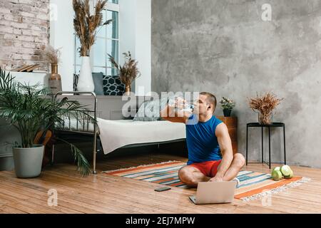 The young woman goes in for sports at home. Cheerful sportswoman with black hair grinking water on the floor at home,  watches a movie and studies fro Stock Photo