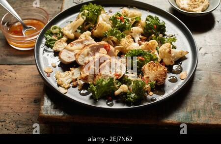 High angle of pieces of grilled chicken with kale and cauliflower served on plate on rustic lumber board near bowls with chili sauce and hummus Stock Photo