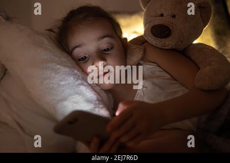 A cute little girl lies on a pillow with a teddy bear and plays on the phone at night in bed. Stock Photo