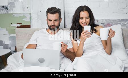 smiling woman chatting on smartphone near man using laptop in bed Stock Photo