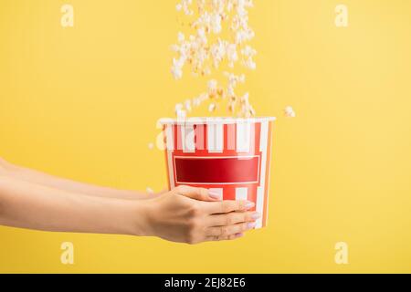 tasty popcorn falling into striped bucket in female hands isolated on yellow, cinema concept Stock Photo