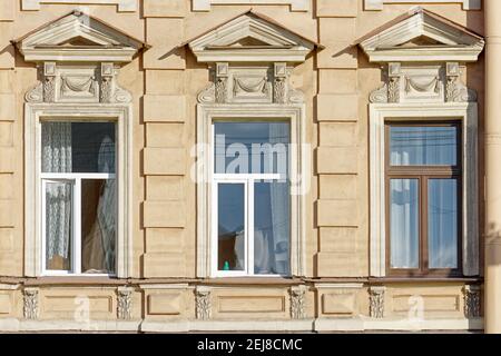 Three rectangular windows with a triangular visor and stucco moldings against the background of an old plastered yellow wall. From a series of windows Stock Photo