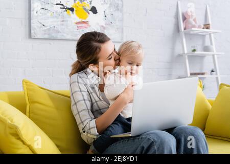 young freelancer kissing little kid while sitting on yellow sofa with laptop Stock Photo