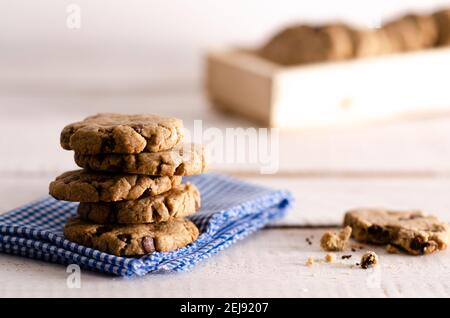 Vanilla and chocolate cookies stacked on a white and blue napkin, a bitten cookie and cookies in a wooden box, on a white wooden backdrop. Stock Photo
