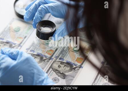 Hands in rubber gloves holding magnifying glass over dollar bills closeup Stock Photo
