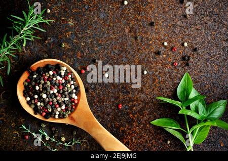 Black, white, green, and pink peppercorns in a wooden spoon, with basil, rosemary, and thyme, on a dark backdrop. Stock Photo