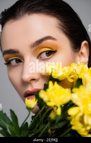 young brunette woman with makeup near blooming yellow flowers on blurred foreground isolated on grey Stock Photo