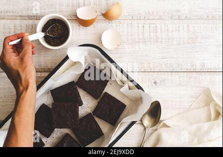Brownies in a white tray with baking paper, egg shells, a hand holding a spoon, and a white napkin and a spoon, on a white wooden backdrop. Stock Photo