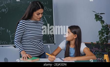 mother standing near upset daughter writing on notepad while doing homework Stock Photo