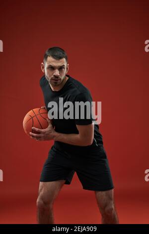 Waist up portrait of adorable strong sportsman in black sportswear holding basketball with both hands isolated on red background Stock Photo