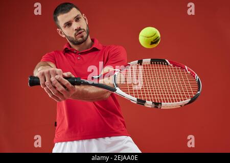 Waist up portrait of handsome Caucasian bearded male in red shirt holding racket in arms while training forehand grip technique on indoor court Stock Photo