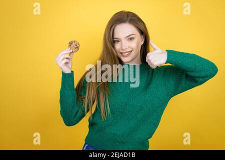 Young beautiful woman eating chocolate cookie over yellow background success sign doing positive gesture with hand, thumb up smiling and happy. cheerf Stock Photo