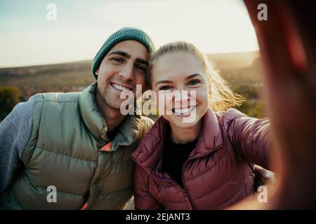 Caucasian husband and wife taking selfie standing on top of mountain smiling while sun sets behind Stock Photo
