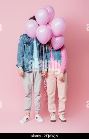 Teen couple holding hands while balloons covering faces on pink background Stock Photo