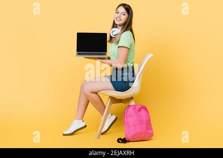 Smiling girl with headphones holding laptop near backpack on yellow background Stock Photo
