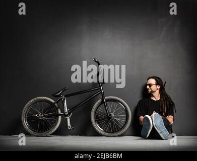 Young smiling man sitting next to his BMX bike. Dreadlocks and sunglasses. Stock Photo