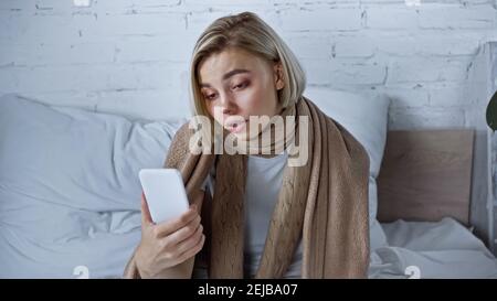 upset sick woman holding smartphone while sitting under warm blanket in bedroom Stock Photo
