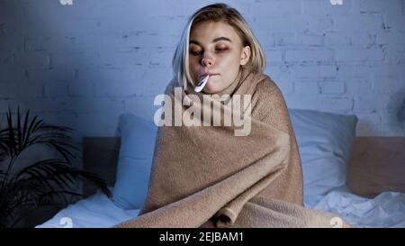 ill woman, wrapped in warm blanket, measuring temperature in bedroom Stock Photo