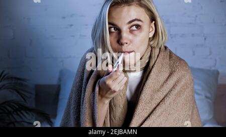 sick woman, wrapped in warm blanket, measuring temperature in bedroom Stock Photo