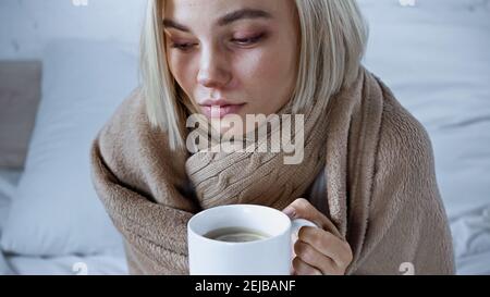 diseased woman, covered with warm blanket, holding cup of tea in bedroom Stock Photo