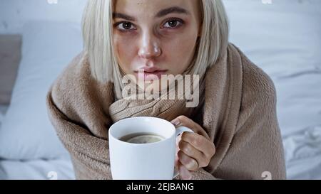 sick woman, wrapped in blanket, holding cup of warm tea in bedroom Stock Photo