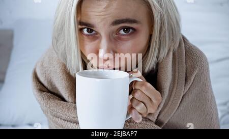 ill woman looking at camera while drinking warm beverage in bedroom Stock Photo