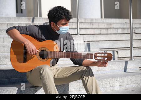 Latin young man wearing protective face mask tuning his guitar sitting on stairs. Playing guitar outdoors. Stock Photo