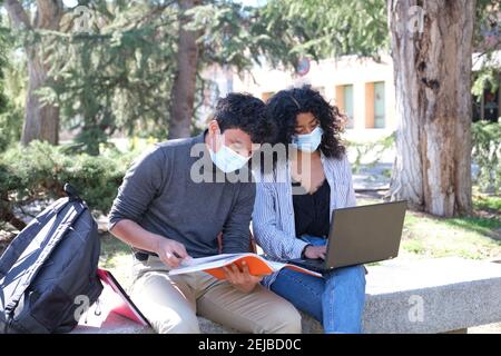 Two latin students wearing protective face mask studying together sitting on a bench outdoor. New normal at university campus. Stock Photo