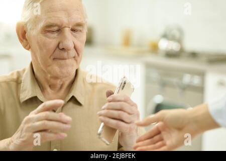Cropped photo of elderly man looking thoughtful while holding a smartphone in his kitchen Stock Photo