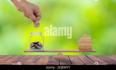 Hands that are putting coins into money saving bottles and model wooden houses on wooden scales in savings ideas for buying a new home or real estate. Stock Photo