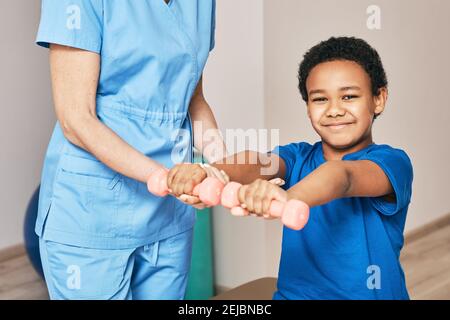 Physiotherapist working with child patient in a rehab clinic. Boy raises dumbbells, strengthening arm muscles and developing joints Stock Photo