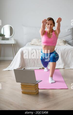 Young woman performs lunges under the guidance of an online trainer in front of a laptop monitor. Online workout at home. Healthy lifestyle concept. Stock Photo