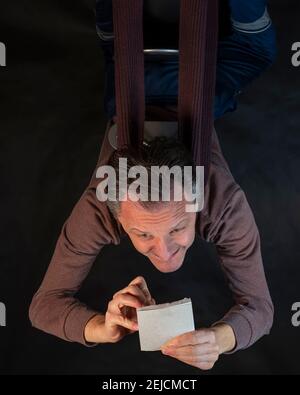 Middle-aged man writes notes on a small notepad sitting upside down with funny expression Stock Photo