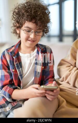 Portrait of cute latin boy wearing glasses smiling while using smartphone, sitting at home. Technology, childhood, homeschooling concept Stock Photo