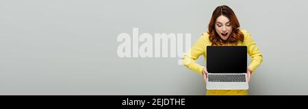 joyful woman standing with open mouth while holding laptop with blank screen on grey, banner Stock Photo