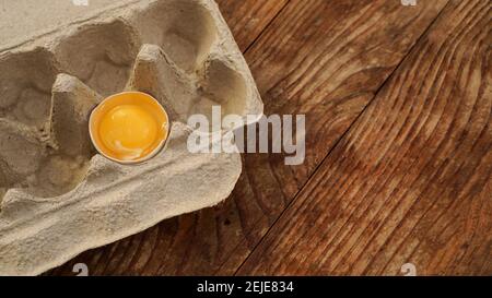 One broken egg in a cardboard egg tray. Half egg with yolk in an empty box on wooden background Stock Photo