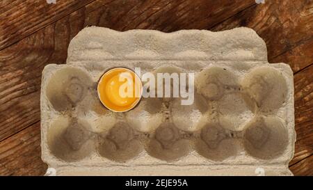 One broken egg in a cardboard egg tray. Half egg with yolk in an empty box on wooden background Stock Photo