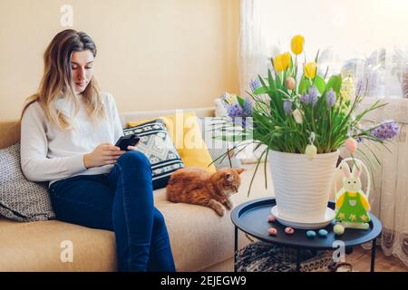Easter home decor. Woman and cat relax on couch. Spring blooming flowers in pot decorated with eggs and bunny. Yellow hyacinths, tulips, muscari in bl Stock Photo
