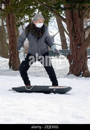 An active Chinese American woman in her 50s sleds down a snowy hill on a cold winter. In a park in Flushing, Queens, New York City. Stock Photo