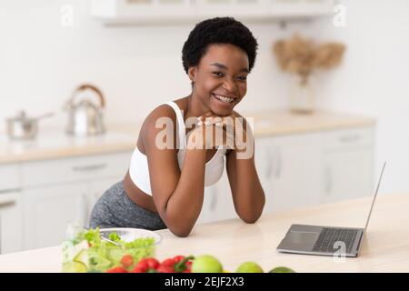 African Woman Using Laptop Browsing Recipes For Weight-Loss In Kitchen Stock Photo