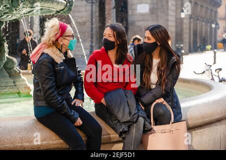 Three female tourists in sunny mediterranean city resting at a fountain, wearing protective face masks. Concept: tourism during covid-19 pandemic Stock Photo