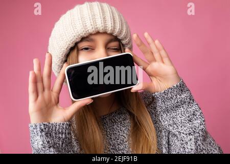 Closeup photo of winking Beautiful young blonde woman wearing casual grey sweater and beige hat isolated over pink background holding in hand and Stock Photo