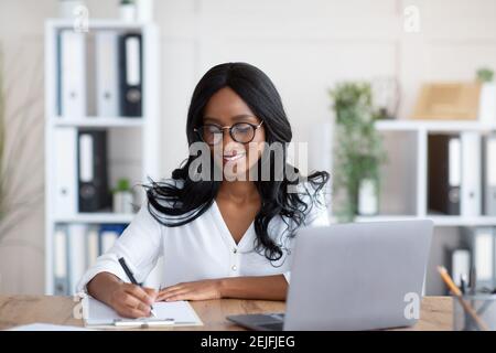 Positive African American female entrepreneur writing down info during online business meeting at office Stock Photo