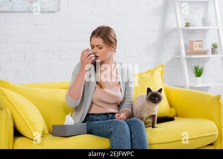 Woman with allergy using inhaler near furry siamese cat on sofa Stock Photo